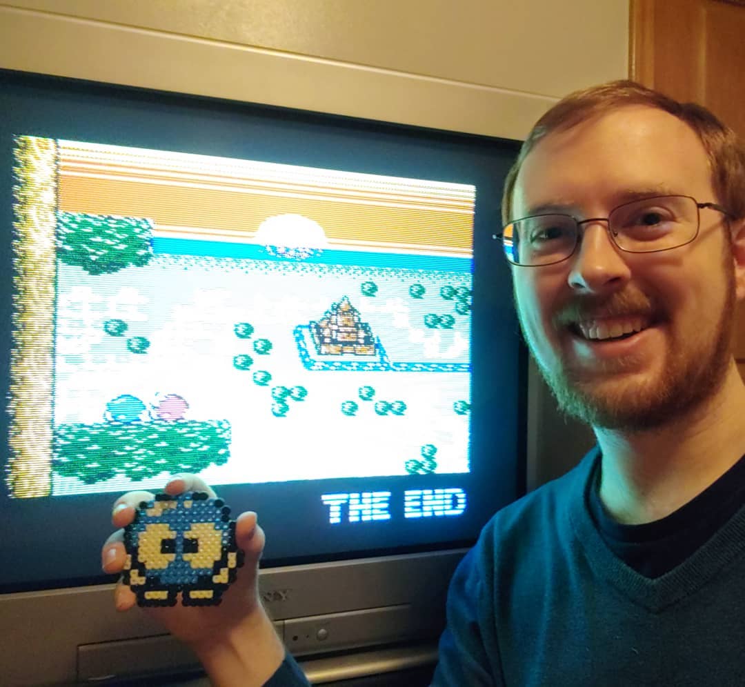 A photo of Tylor holding a perler bead reconstruction of the sprite of Lolo in front of a CRT displaying the end screen of Adventures of Lolo 3 for the NES