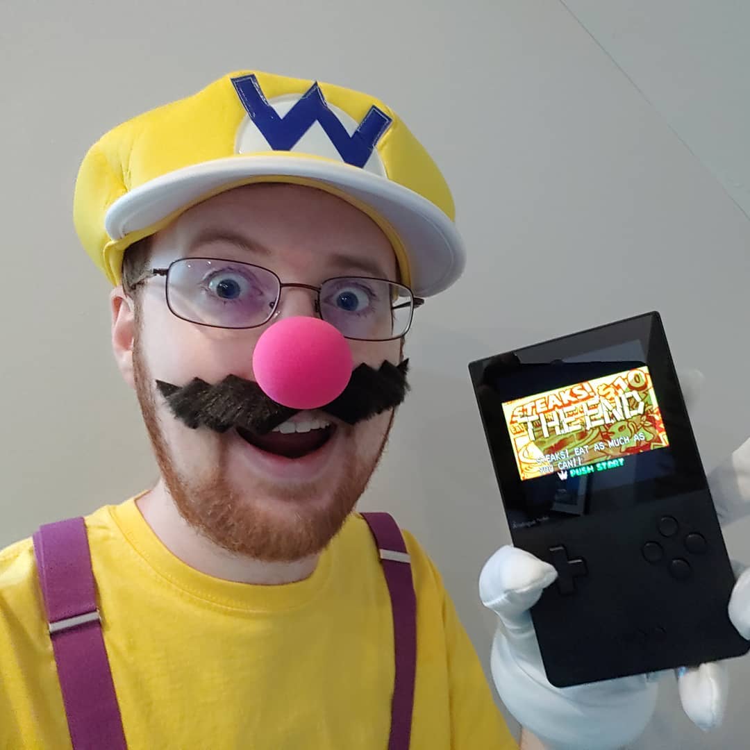 A photo of Tylor dressed in a Wario Costume, holding an Analogue Pocket that displays the end screen of Wario Land 4